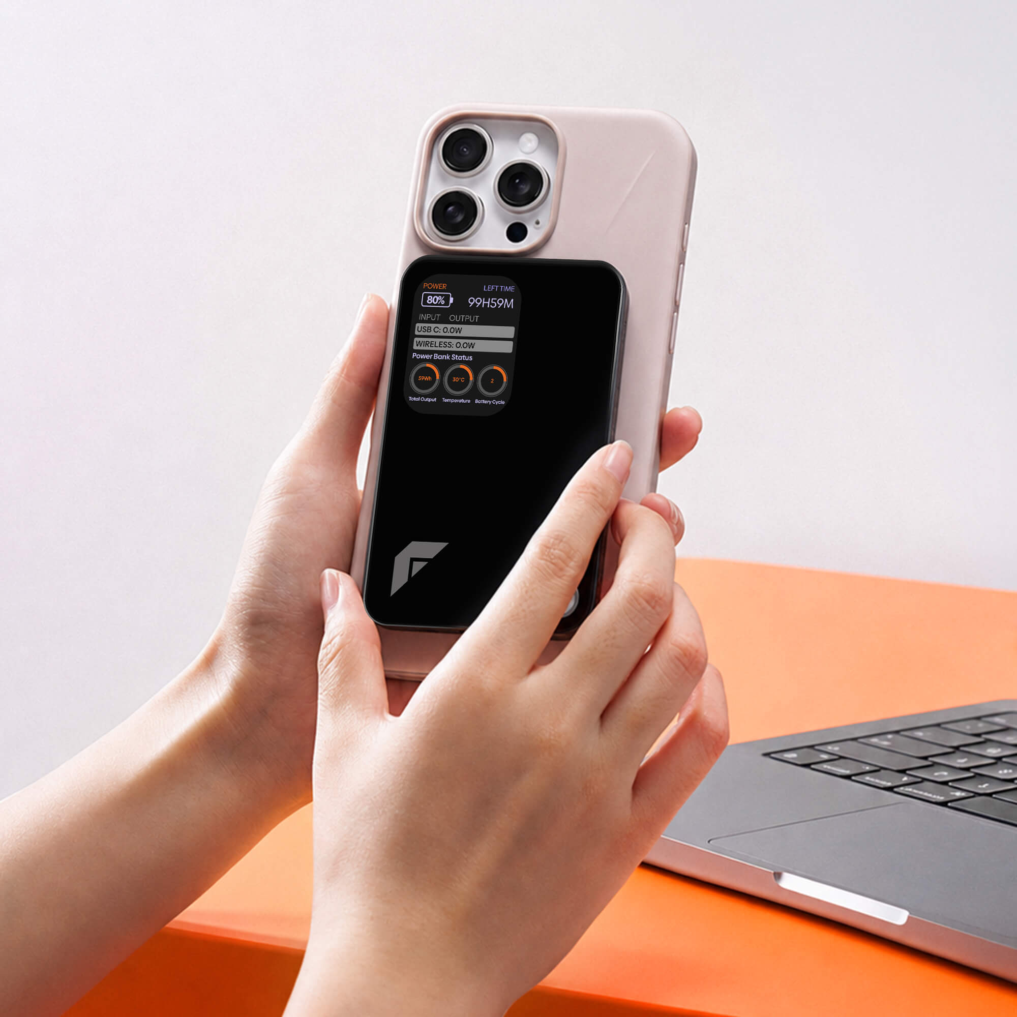 A person holds a light pink phone case with a built-in black magnetic power bank module. The phone inside is visible through a transparent window, and a laptop sits on an orange desk in the background.