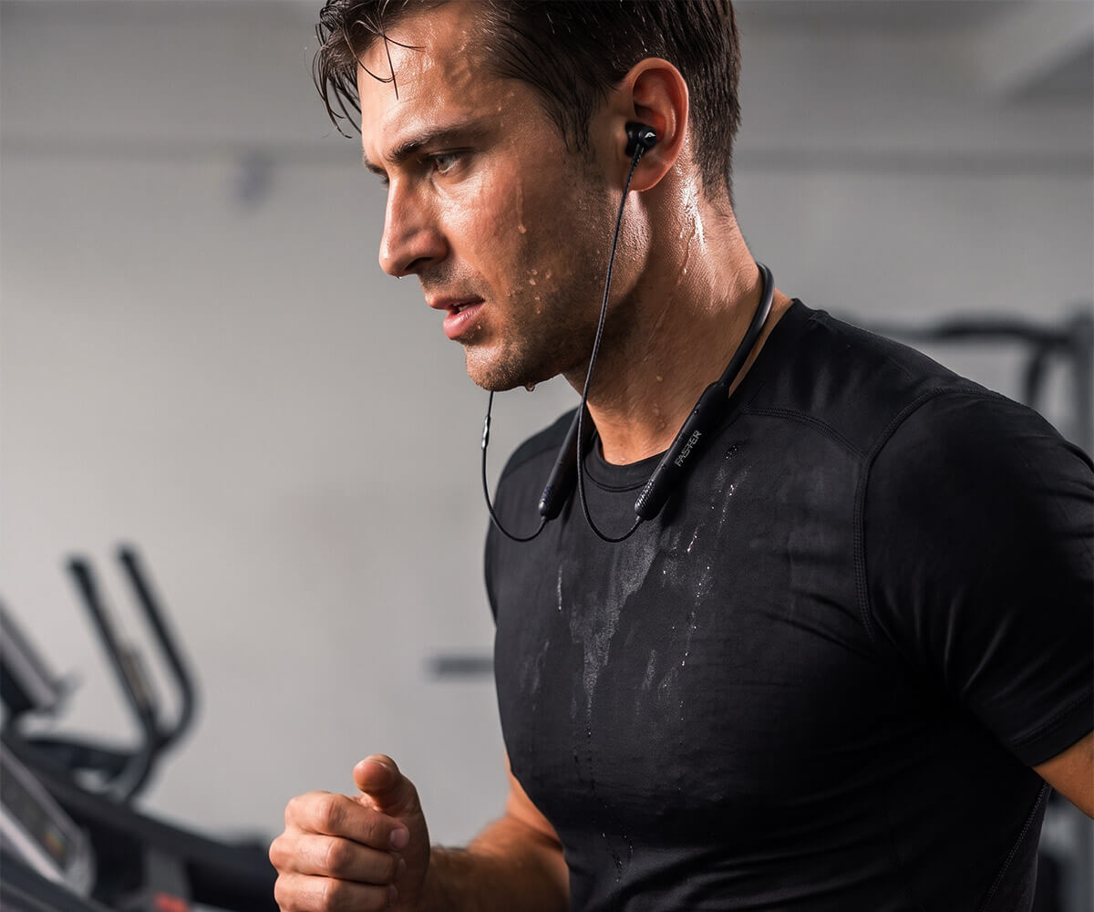A man in a black athletic t-shirt running in a gym while wearing a black neckband with wireless neckband.