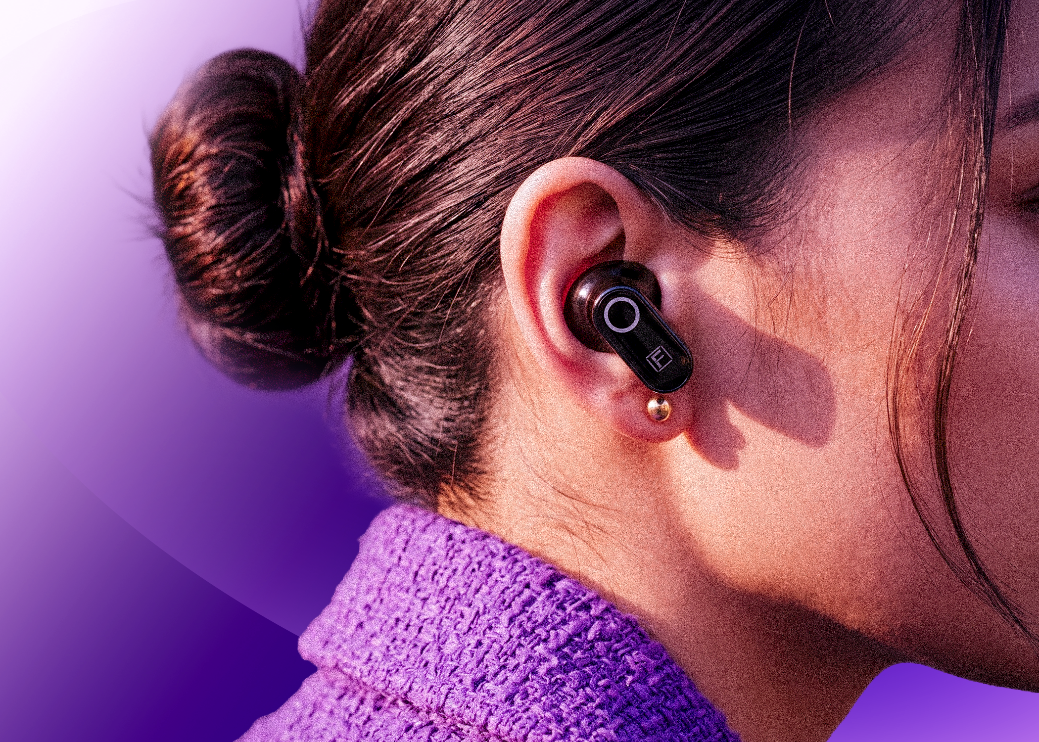 A close-up side profile of a person wearing a black in-ear wireless earbud, with dark hair in a bun and a textured purple top against a purple gradient background.