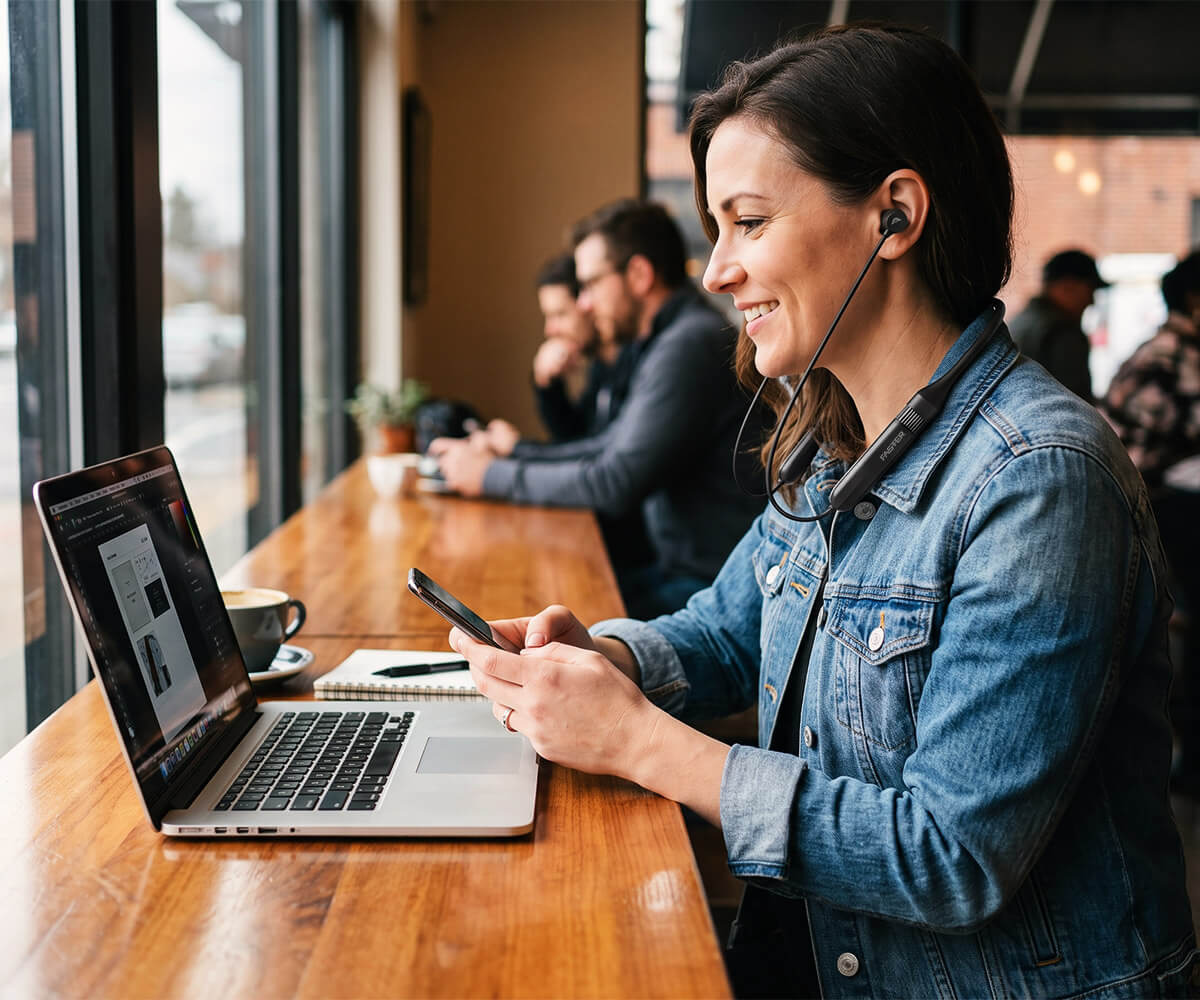 A woman in a blue denim jacket sits at a wooden table in a cafe, wearing a headset and looking at her phone next to an open laptop.