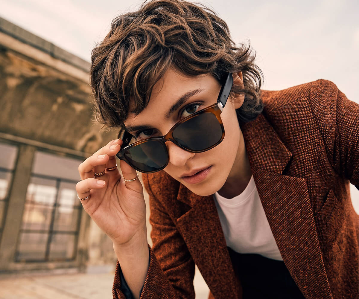 A close-up lifestyle shot of a person with wavy brown hair wearing tortoiseshell smart glasses and a brown tweed blazer over a white t-shirt, holding the sunglasses in front of their face against an outdoor architectural background.